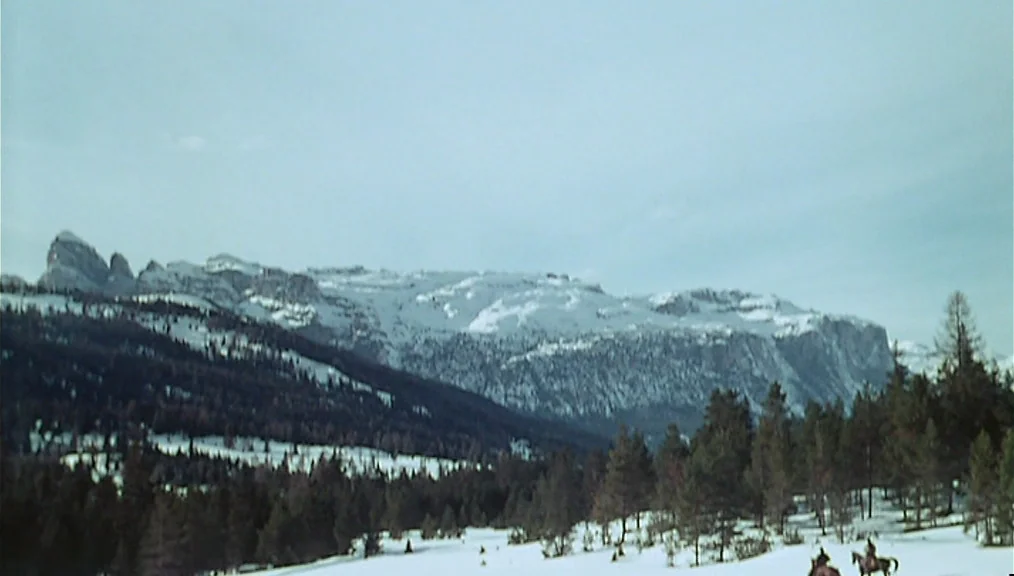 The Great Silence (1968) movie still: wide — A vast, snowy landscape sits beneath high, jagged, snow-capped mountain peaks. A dense pine forest…