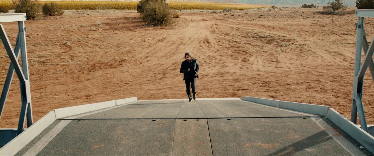 The Last Stand (2013) movie still: wide — An arid desert landscape features a concrete overpass bridge stretching into the distance. A man in a dark…