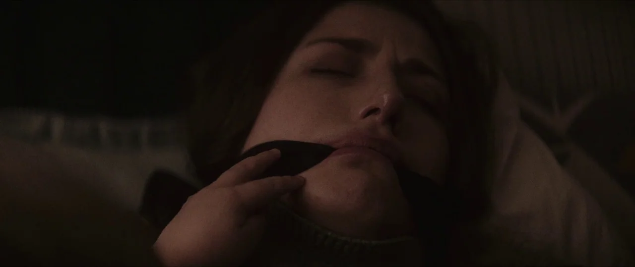 Anything For Jackson (2020) movie still: closeup — A dimly lit interior space features a woman lying on a soft surface. She has a dark fabric gag…