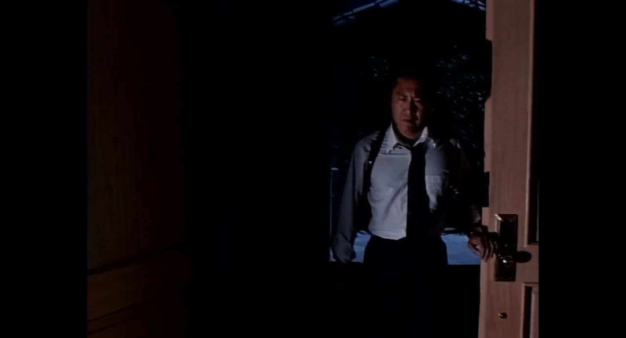 American Yakuza (1993) movie still: medium — A dark interior doorway frames a man standing at the threshold. The man looks forward while gripping…