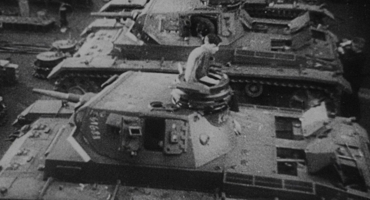Darkest Hour (2017) movie still: wide — A military motor pool contains multiple parked armored tanks in a paved lot. A soldier stands partially…