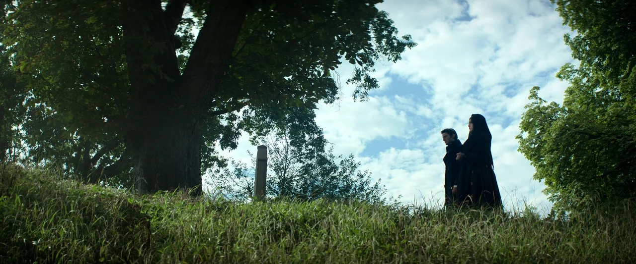 The Count of Monte-Cristo (2024) movie still: wide — A grassy hill holds a large dark tree and a stone headstone. A man and a woman in black…