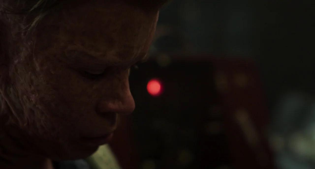 The Little Stranger (2018) movie still: closeup — A dimly lit, shadowed space contains a figure with visible facial scarring. The individual looks…