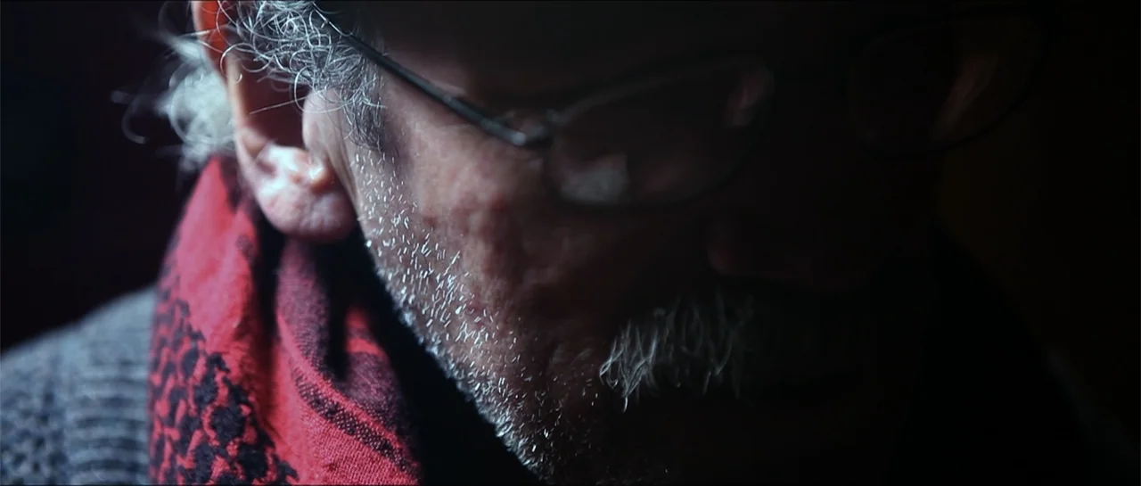 Garnet’s Gold (2014) movie still: closeup — A dimly lit interior space is obscured by heavy shadow. An elderly man with a grey mustache and glasses…