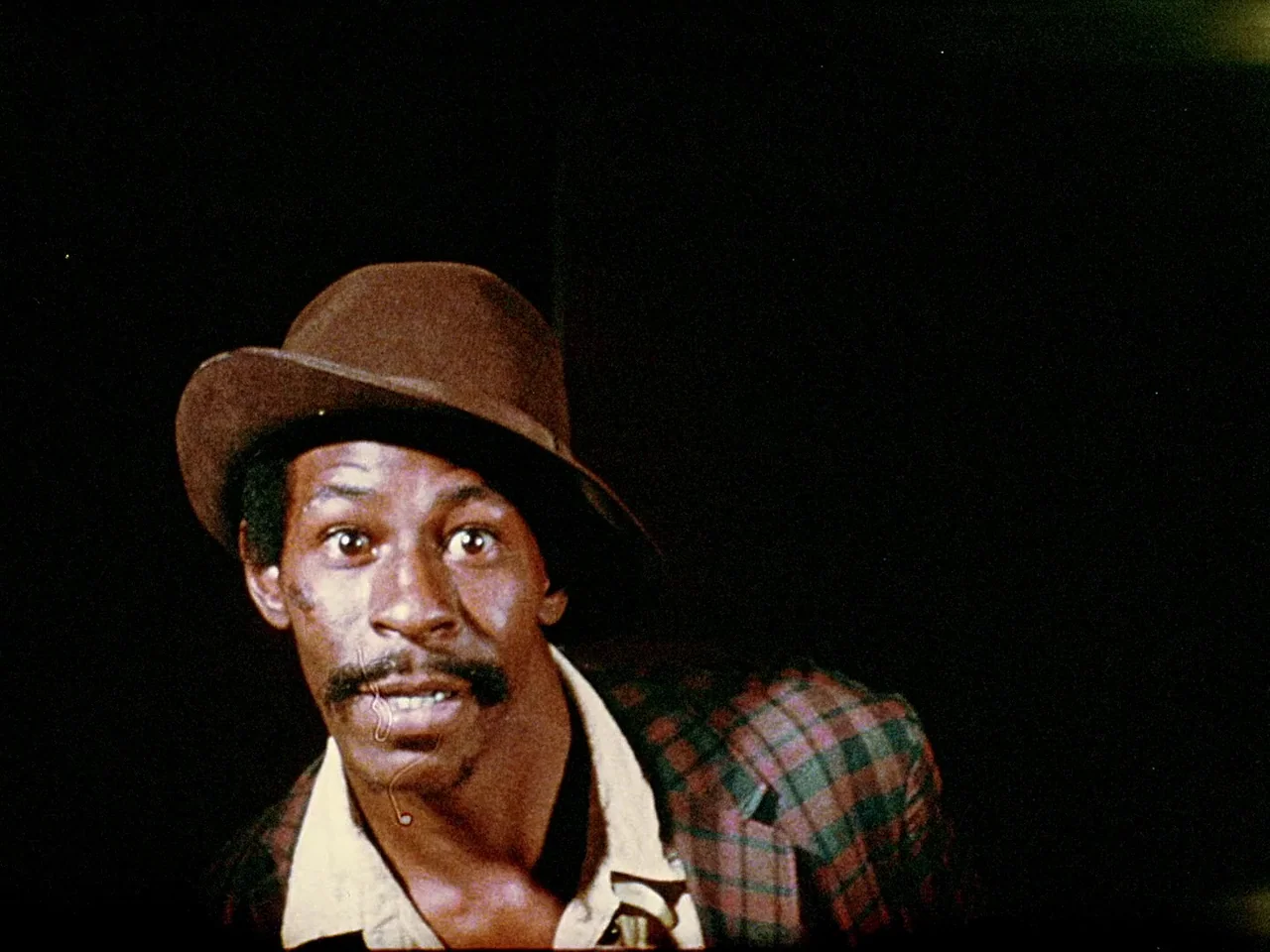 Space is the Place (1974) movie still: closeup — A man stands against a pitch black background in a dimly lit environment. He looks forward with…