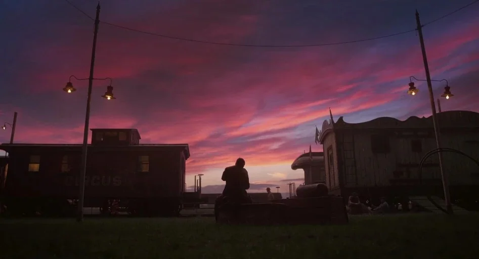 Dumbo (2019) movie still: wide — A circus train yard features wooden wagons and railcars under a dramatic sunset. A lone man sits on a low crate…