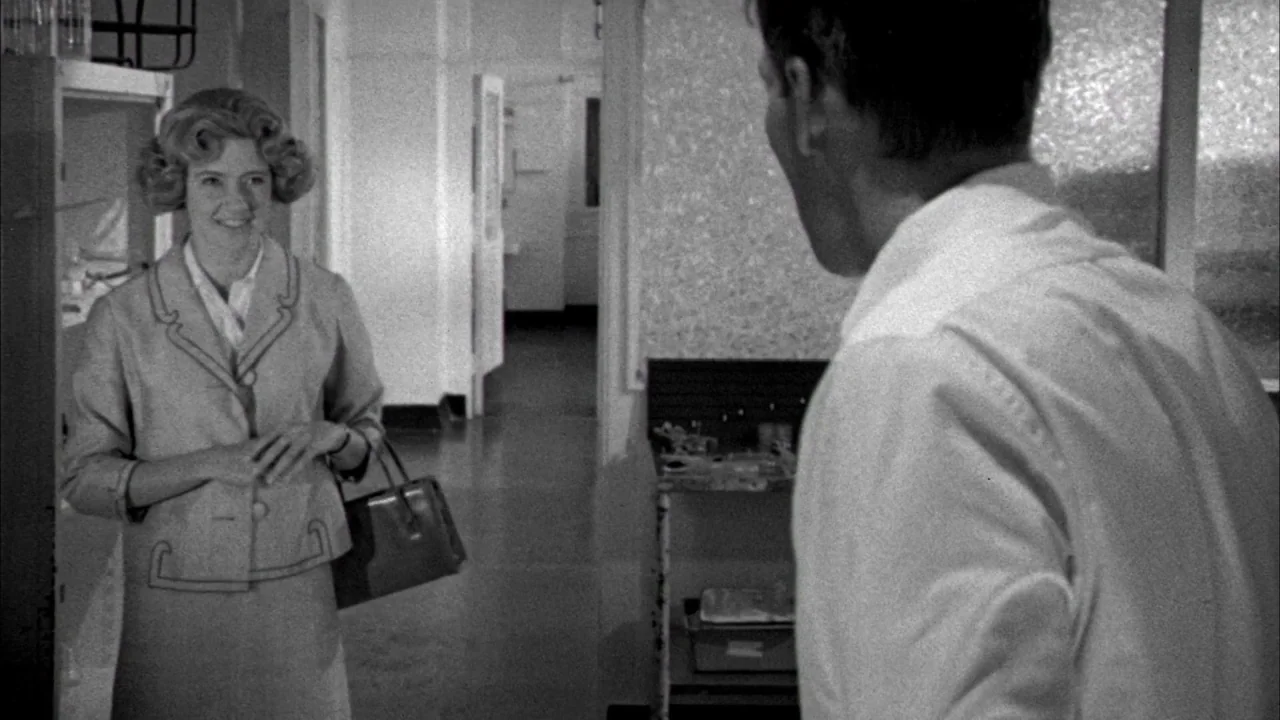 Poison movie still: medium — A sterile hallway within a medical facility features clinical white walls and tiled floors. A woman in a suit stands…