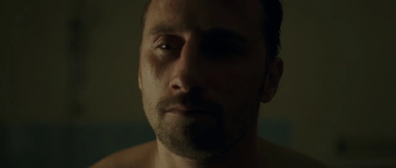 Bullhead (2011) movie still: closeup — A dimly lit, shadowed bathroom interior with muted green tones and minimal architectural detail. A man stands…