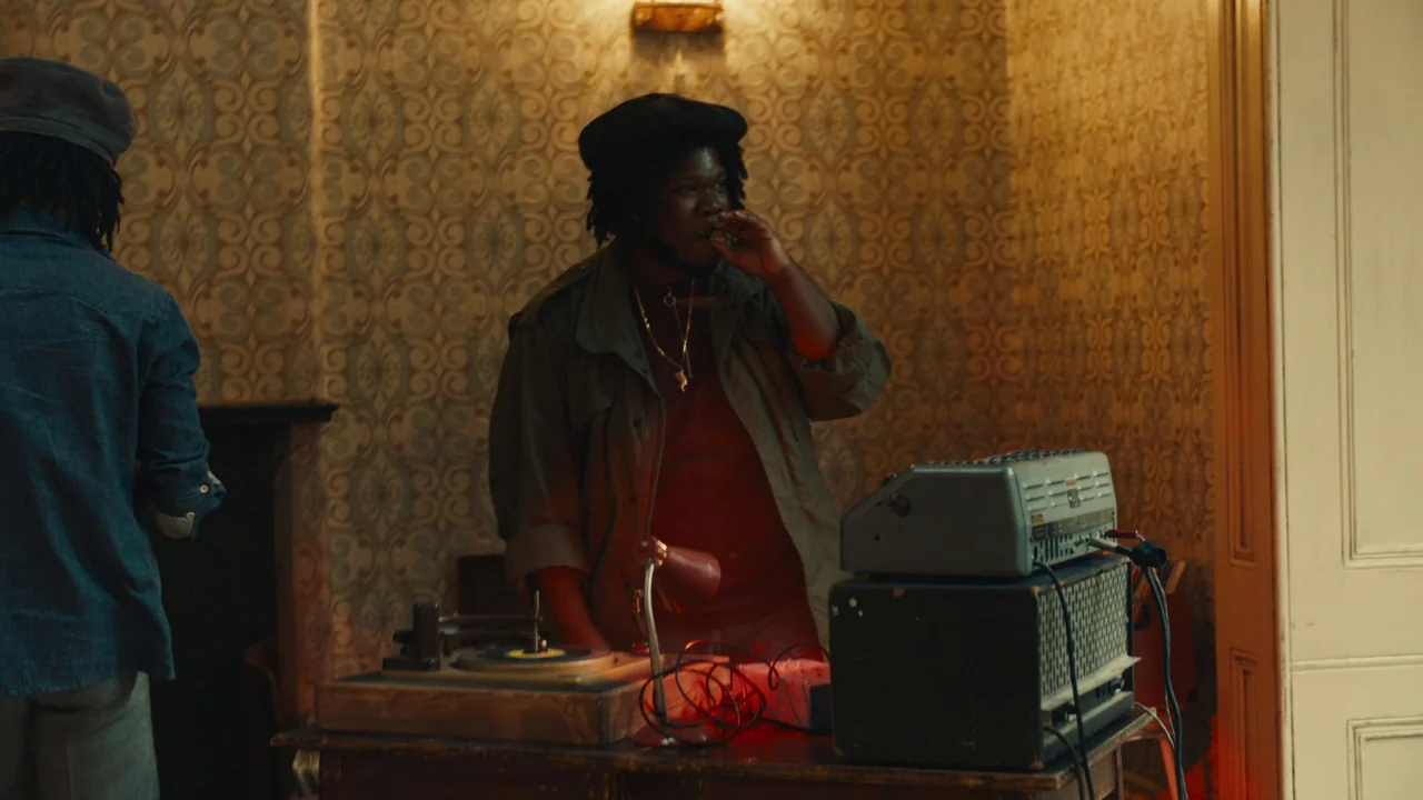 Lover’s Rock (2020) movie still: medium — A dimly lit room with patterned wallpaper features an audio setup on a wooden table. A man in an olive…