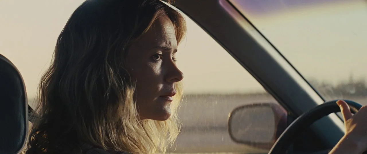 Mud (2012) movie still: closeup — The interior of a moving vehicle during a bright golden hour. A woman sits behind the steering wheel while looking…