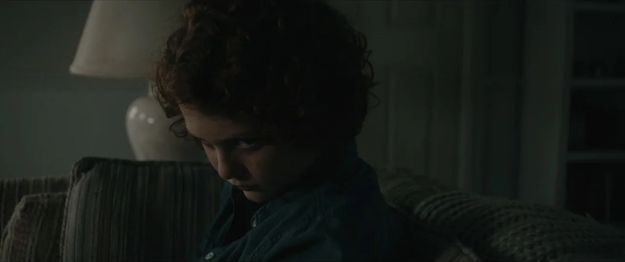 Possessions (2024) movie still: closeup — A dark, dimly lit living room features a child seated on a fabric couch. A boy with curly hair looks…