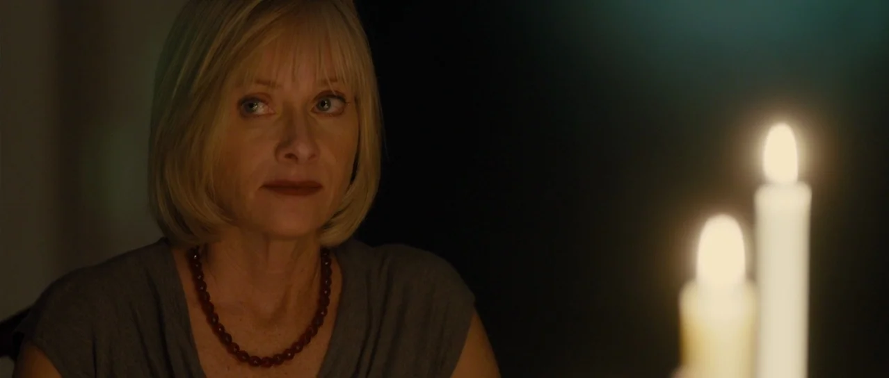 Road Games (2015) movie still: closeup — A woman sits in a dark room illuminated by the glow of candles. She looks toward the side while sitting at…