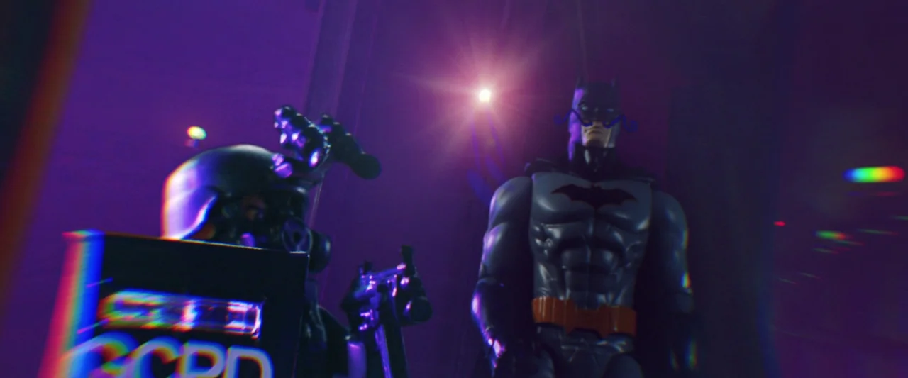 The People’s Joker (2022) movie still: medium — A dark space features a plastic Batman action figure beside an armored tactical officer. The…