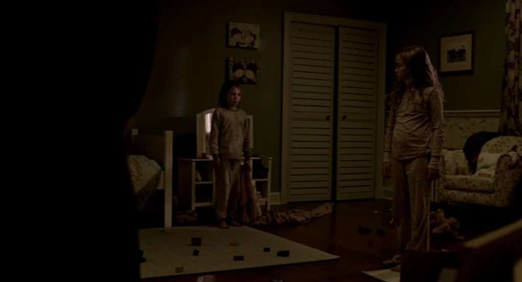 Mama (2013) movie still: wide — A dimly lit child's room features dark green walls, patterned wallpaper, and wooden flooring. Two young girls stand…