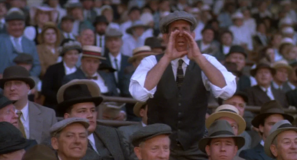 Eight Men Out (1988) movie still: medium — A dense crowd of men in hats sits in outdoor stadium bleachers. One man in the foreground cups his hands…