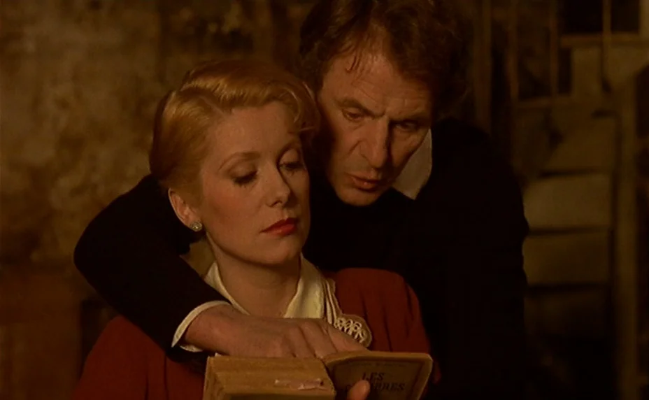 The Last Metro (1980) movie still: medium — A dimly lit room with muted walls and a staircase visible in the background. A man stands behind a woman…