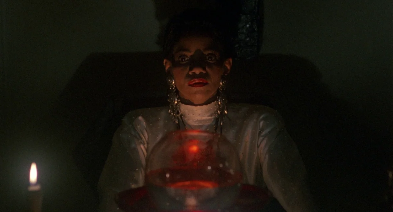 Def By Temptation (1990) movie still: medium — A dark space lit only by a candle and a glowing red orb. A woman looks forward with an intense…