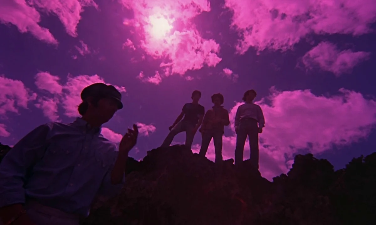 Help! (1965) movie still: wide — Four men stand atop a dark, jagged rock formation against a vibrant purple sky. The camera looks up from a low…