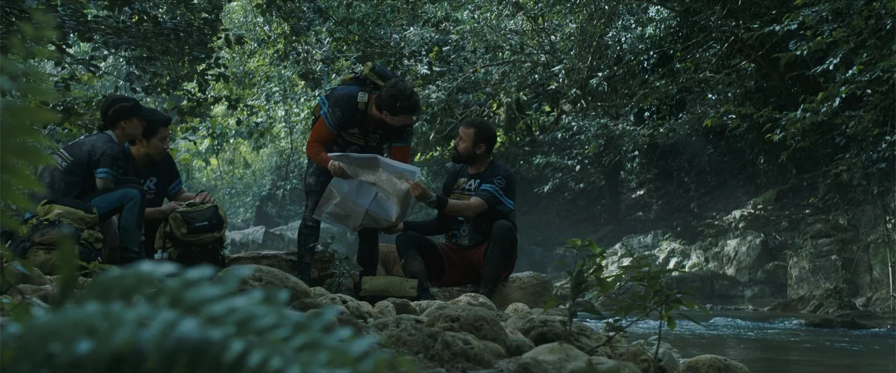 Arthur the King (2024) movie still: wide — A group of four athletes in athletic gear navigate through a dense, green jungle setting. They crouch…