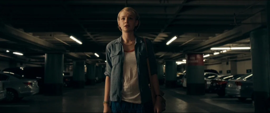 Drive (2011) movie still: medium — A subterranean parking garage with rows of parked vehicles and concrete support pillars. A woman stands centered…