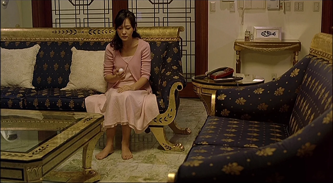 3-Iron (2004) movie still: medium — A formal interior features ornate dark blue sofas with gold trim and wood accents. A woman sits on the sofa…
