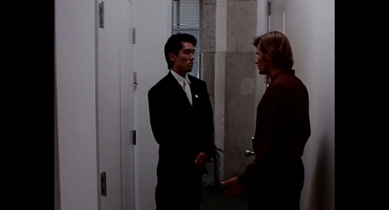American Yakuza (1993) movie still: medium — A narrow office hallway features white walls, a closed white door, and gray concrete accents. Two men…