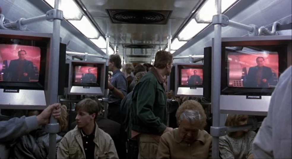 Total Recall (1990) movie still: medium — A crowded modern transit car interior is illuminated by rows of overhead fluorescent lights. Passengers…