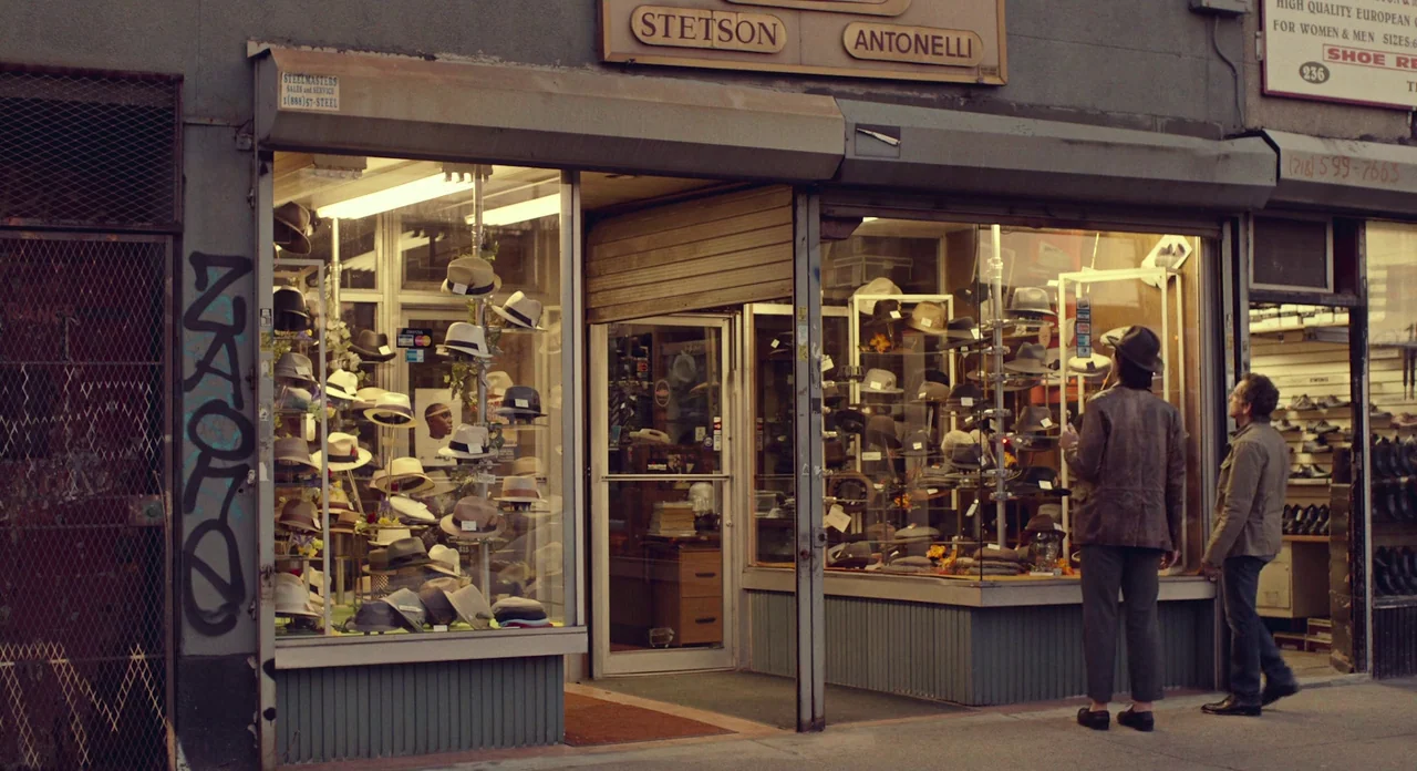 While We’re Young (2014) movie still: wide — A street-level view of a hat shop with large glass windows displaying multiple fedoras. Two men stand…