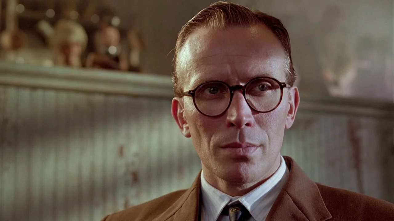 Naked Lunch (1991) movie still: closeup — A man in a blazer stands before a wood-paneled wall inside a public house. He stares directly into the…