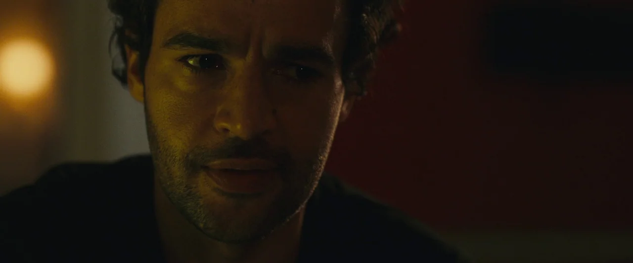 Sanctuary (2023) movie still: closeup — A dimly lit interior space with an indistinct dark background. A man with dark wavy hair looks toward the…