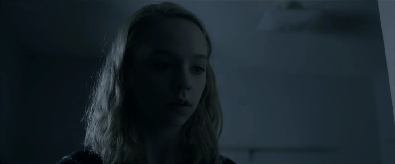 The Hollow Child (2018) movie still: closeup — A dimly lit interior room at night features a young woman in shadow. She stands facing off-screen…
