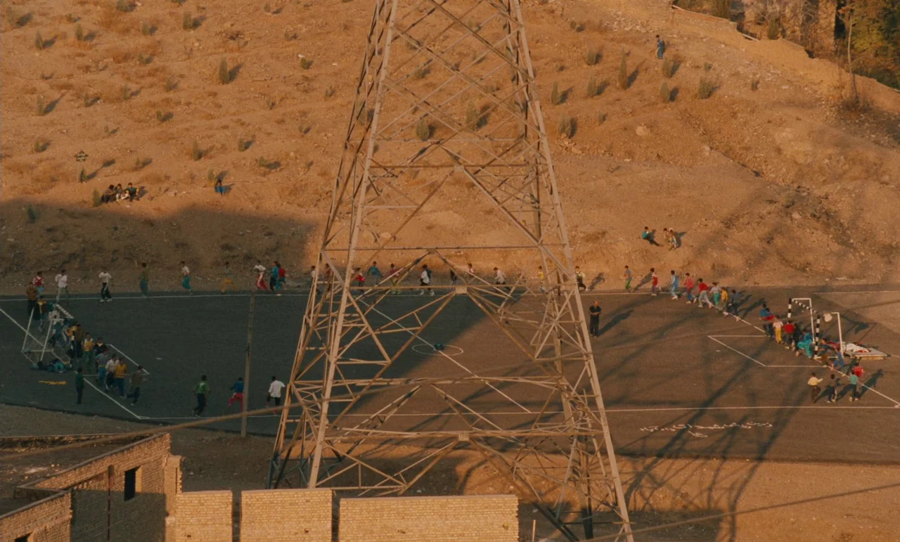 Taste of Cherry (1997) movie still: wide — A large metal electrical tower stands centrally in a flat, arid sports field. Many children are scattered…