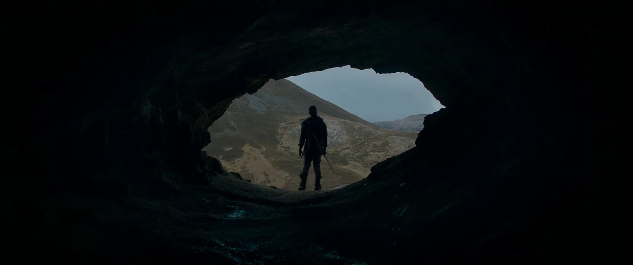 Out of Darkness (2022) movie still: wide — A dark, uneven cave opening frames a view of a rolling, grassy mountain slope. A lone person stands at…