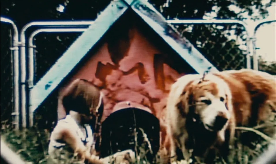 Blue Valentine (2010) movie still: medium — A wooden doghouse stands in a backyard enclosure with chain-link fencing behind it. A young child with…