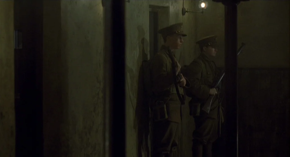 The Wind That Shakes the Barley (2006) movie still: medium — A dark military bunker with rough concrete walls and low lighting levels. Two soldiers…