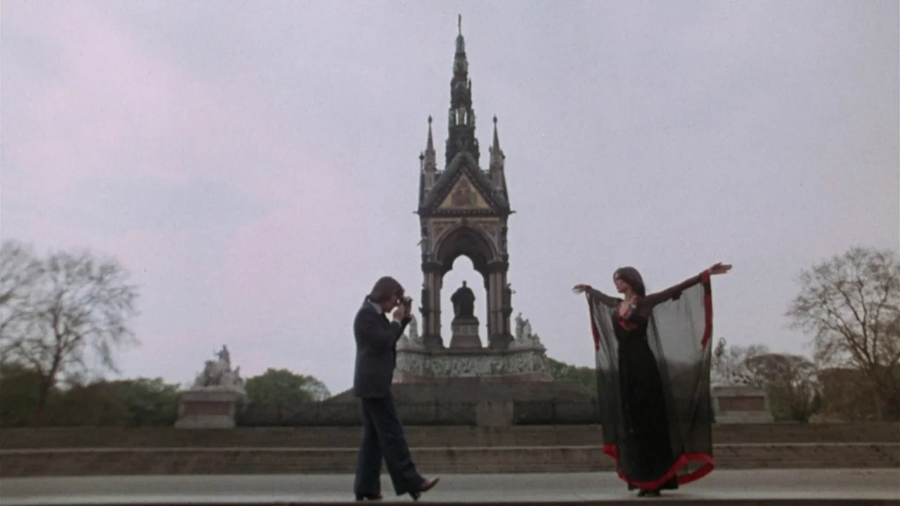 The Sentinel (1977) movie still: wide — A stone gothic memorial structure stands against an overcast sky in a public square. A man in a suit takes a…