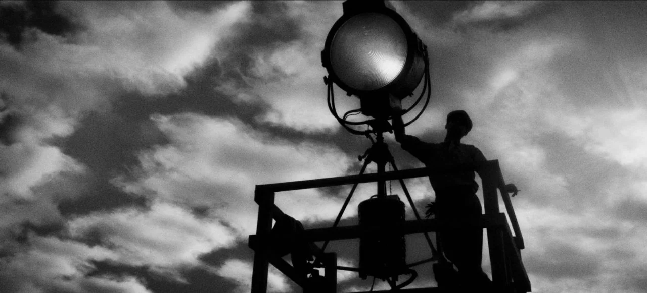Mank (2020) movie still: wide — An industrial metal platform stands against a sky filled with dark, textured storm clouds. A man in silhouette…
