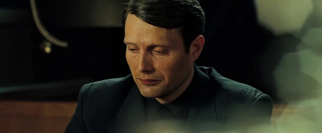 Casino Royale (2006) movie still: closeup — A man in a dark suit sits in a dimly lit office environment. The man looks down with his eyes closed…