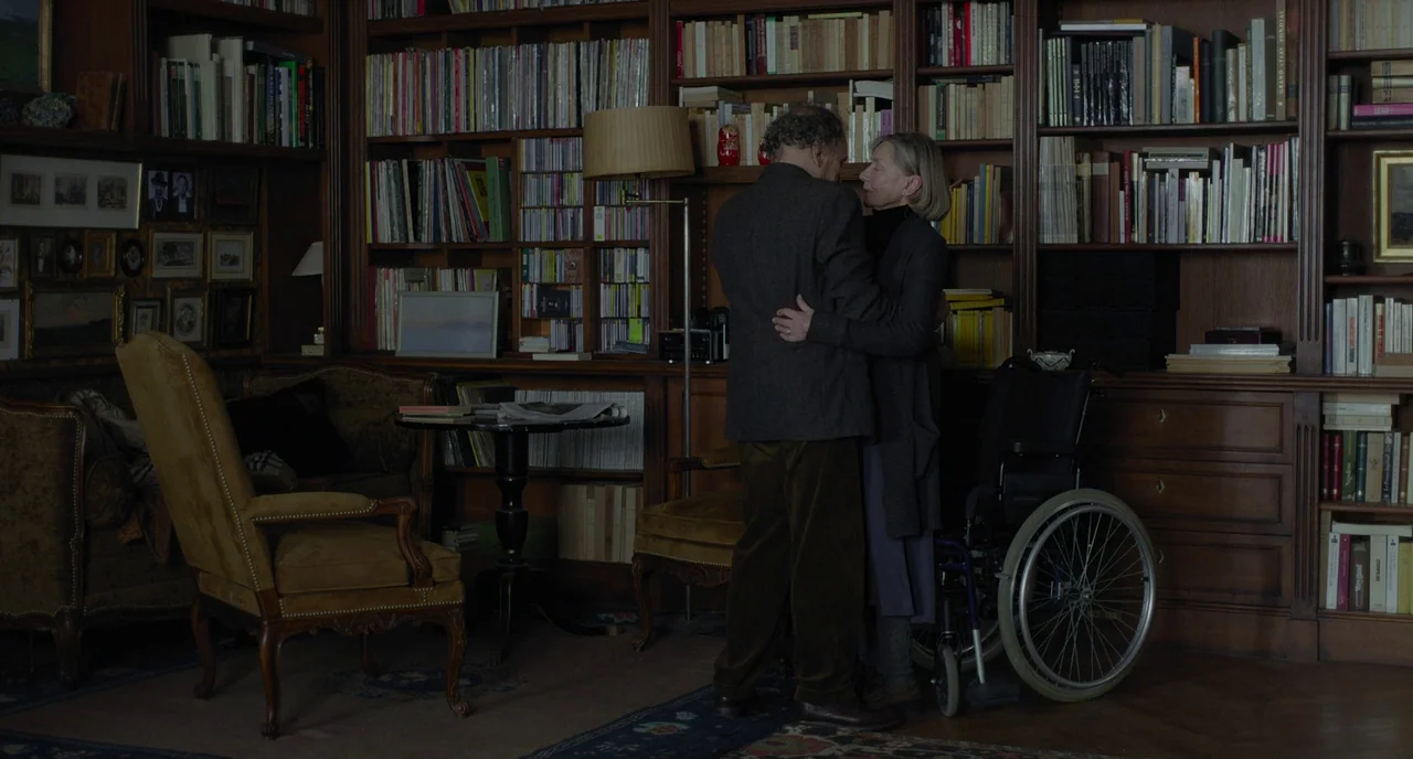 Amour (2012) movie still: fullbody — A dark wood-paneled room filled with extensive floor-to-ceiling bookshelves and framed pictures. An elderly…