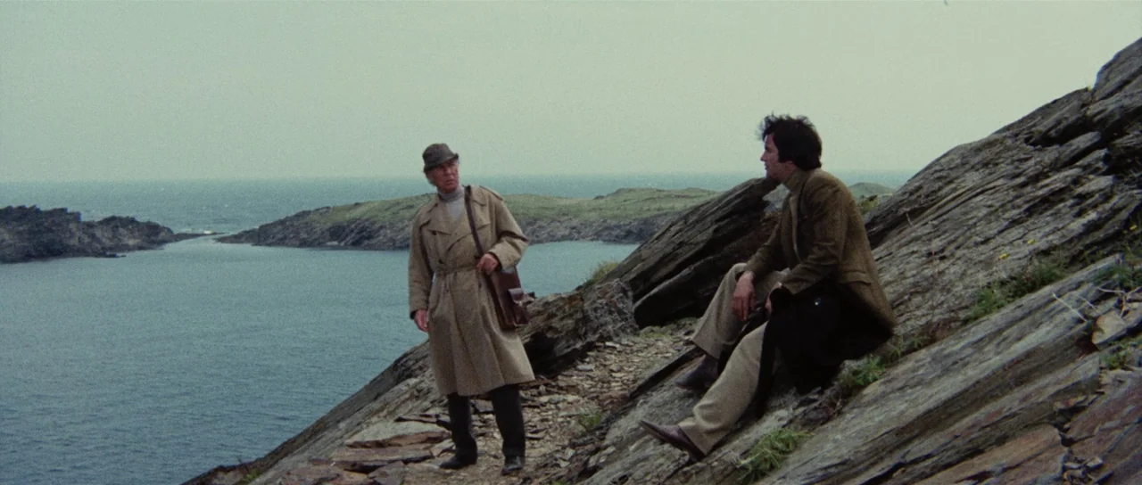 Death Walks On High Heels (1971) movie still: wide — A rugged, rocky coastline overlooks a bay with small islands under an overcast sky. Two men in…