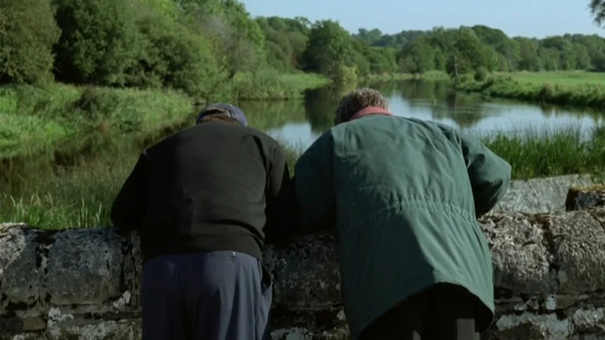 Garage (2007) movie still: medium — Two men stand on a rustic stone bridge overlooking a calm, winding river. Lush green trees and grass banks frame…