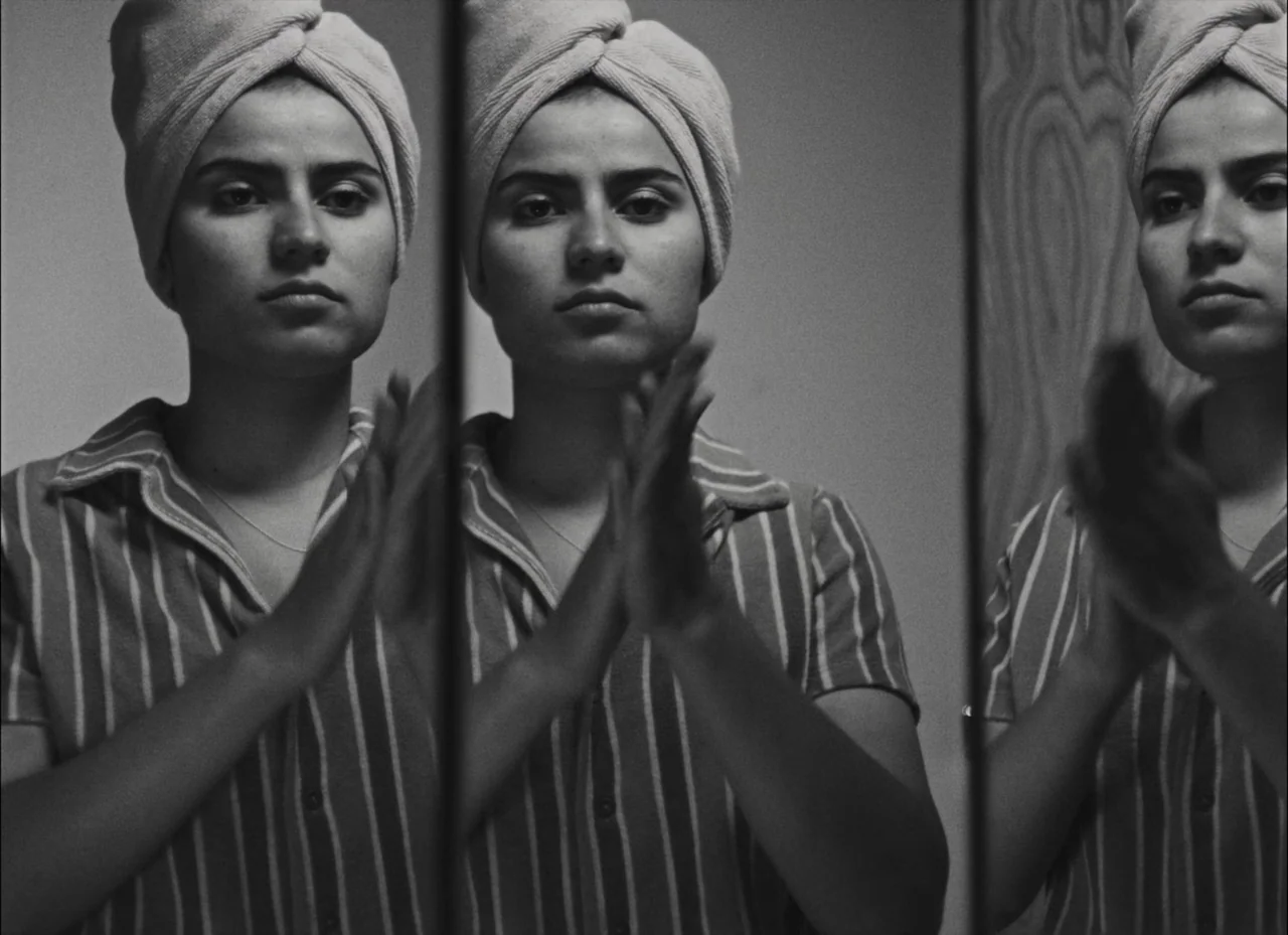 Fremont (2023) movie still: medium — A woman stands in front of a tri-fold mirror in a bathroom setting. She claps her hands together while looking…