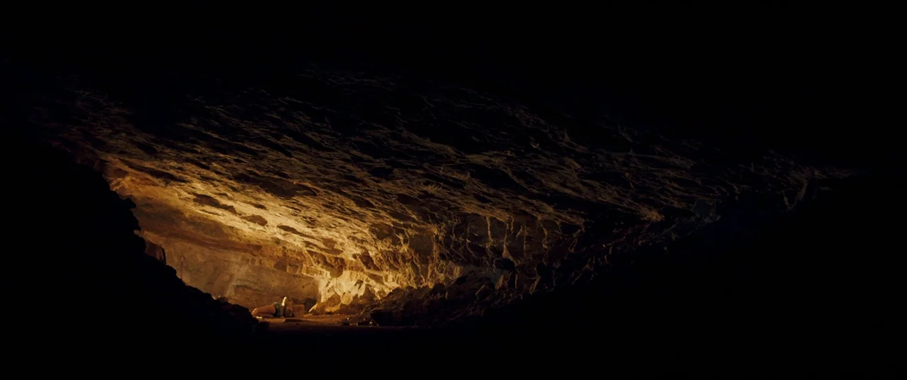 Revenge (2017) movie still: wide — A vast underground cave features jagged rocky walls and a textured stone floor. Harsh warm light catches the top…