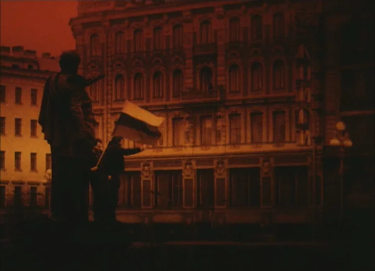 Russian Symphony (1994) movie still: wide — A dark, silhouetted figure stands beside a large statue in a city square. The individual holds a flag in…