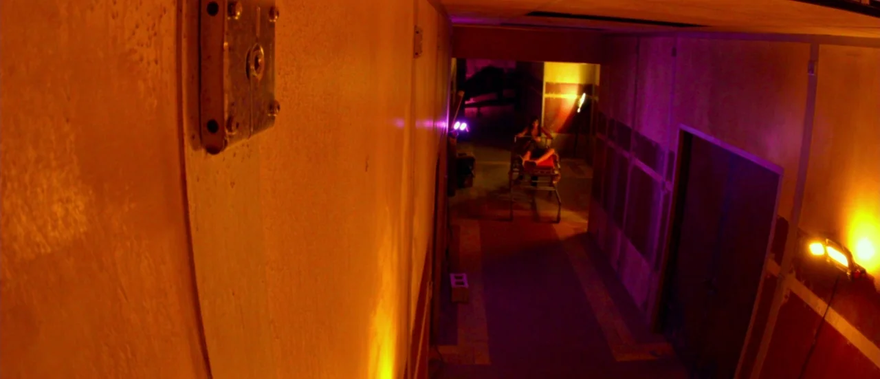 Rupture (2016) movie still: wide — A long interior hallway features concrete walls painted in warm orange and dark red. A single person sits…