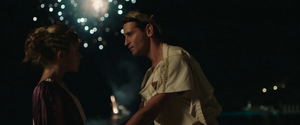 Greed (2019) movie still: medium — A dark outdoor space at night with fireworks bursting in the background. A man in a laurel wreath faces a woman…