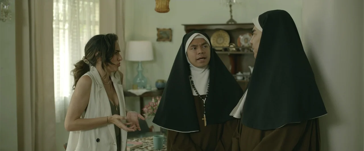 Divine Intervention (2026) movie still: medium — A sunlit dining room features a hutch filled with plates and a table lamp. A woman talks to two…