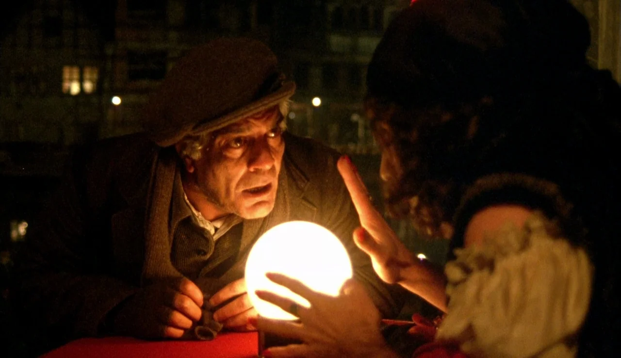 The Rainbow Thief (1990) movie still: medium — A dimly lit room with a man and woman leaning over a table. The man watches as the woman's hands…