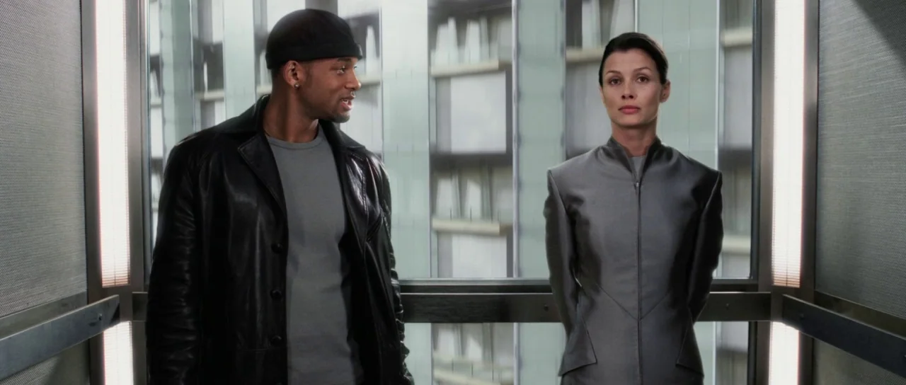I, Robot (2004) movie still: medium — A glass elevator rising past a modern office building with white window frames. A man in a leather jacket…