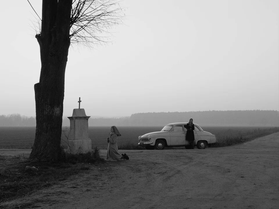 Ida (2013) movie still: wide — A flat, overcast landscape features a dirt road beside a bare tree and shrine. One woman kneels to pray while another…
